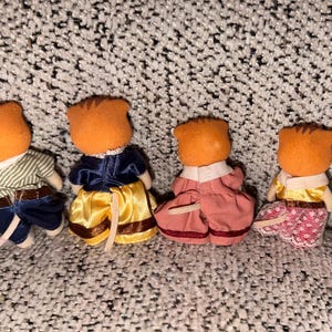 Puede incluir: Cuatro figuras de animales en miniatura con cabezas naranjas y varios atuendos. Uno viste una camisa a rayas y pantalones azules, otro una chaqueta azul y una falda amarilla, un tercero un vestido rosa y el &uacute;ltimo una blusa amarilla y una falda estampada.