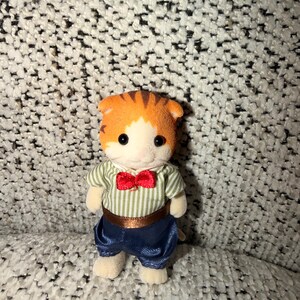 Puede incluir: Una peque&ntilde;a figura de gato naranja y blanco, vestida con una camisa a rayas verdes y blancas, pantalones azules y una pajarita roja. El gato de juguete tiene ojos negros y est&aacute; sobre una superficie texturizada.