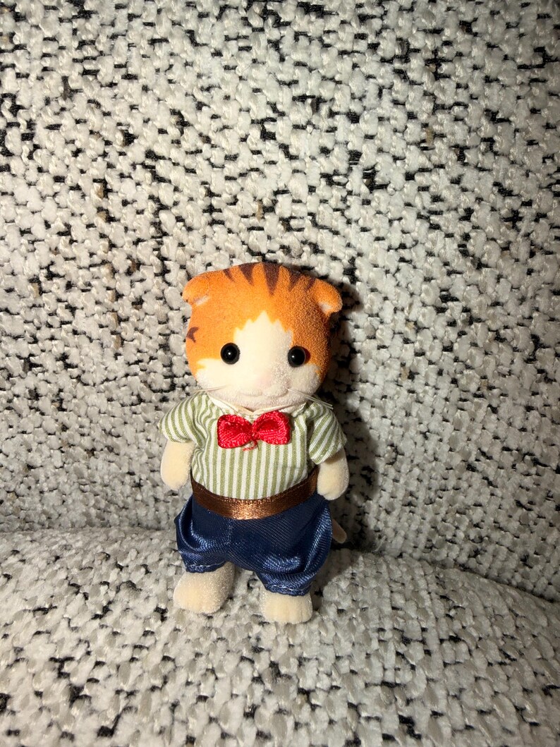 Puede incluir: Una peque&ntilde;a figura de gato naranja y blanco, vestida con una camisa a rayas verdes y blancas, pantalones azules y una pajarita roja. El gato de juguete tiene ojos negros y est&aacute; sobre una superficie texturizada.