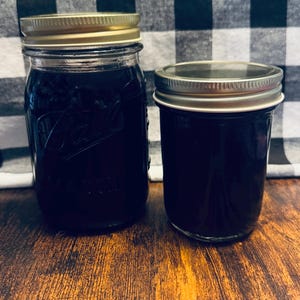 May include: Two glass Mason jars filled with a dark purple liquid, likely preserves or jelly. The jars have gold-colored lids and are set on a wooden surface. The background features a black and white checkered pattern.