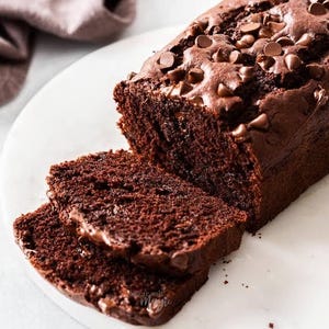 May include: A sliced chocolate loaf cake with chocolate chips on top. The cake is dark brown and sits on a white plate. The slices show a moist interior, perfect for dessert.