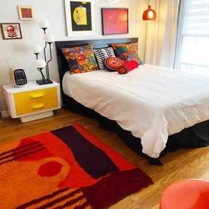 May include: A white bed with a black headboard and a white duvet cover. The bed has colorful pillows and a red and orange rug on the floor. There is a yellow nightstand with two drawers and a lamp with white globes.