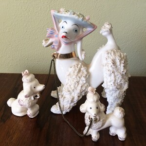 May include: A set of three white porcelain poodle figurines. The largest poodle is wearing a blue and pink hat and a gold chain. The other two poodles are smaller and are also wearing gold chains.