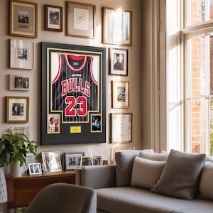 Puede incluir: Una camiseta negra de los Chicago Bulls enmarcada con detalles en rojo y blanco, que muestra el n&uacute;mero 23 y el nombre Jordan. La exhibici&oacute;n incluye dos fotograf&iacute;as peque&ntilde;as y una placa dorada, todo dentro de un marco y paspart&uacute; negros.