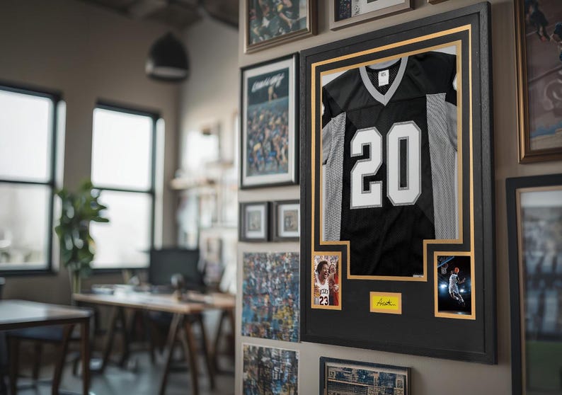 Puede incluir: Una camiseta deportiva en blanco y negro con el n&uacute;mero 20 exhibida en un marco negro grande. La pieza incluye dos fotos rectangulares peque&ntilde;as y una placa rectangular amarilla con una firma, montadas sobre un paspart&uacute; negro con borde dorado.