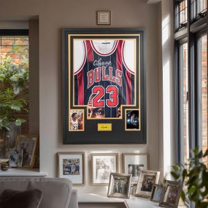 Puede incluir: Una camiseta de baloncesto de los Chicago Bulls enmarcada con el n&uacute;mero 23 en rojo y blanco, con rayas verticales. La exhibici&oacute;n incluye dos fotograf&iacute;as peque&ntilde;as de un jugador de baloncesto y una placa dorada con una firma sobre fondo negro.