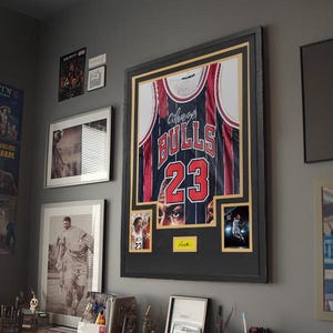 Puede incluir: Una camiseta de los Chicago Bulls enmarcada con el n&uacute;mero 23 en rojo y blanco, exhibida en un marco negro con paspart&uacute; dorado. La pieza incluye dos fotograf&iacute;as peque&ntilde;as insertadas y una placa amarilla, rodeada de otros cuadros.