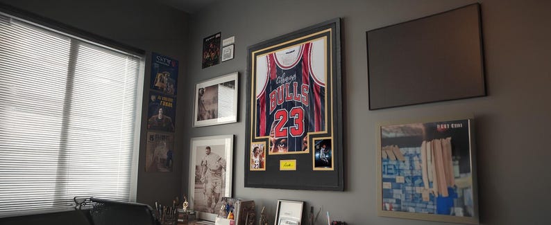 Puede incluir: Una camiseta de los Chicago Bulls enmarcada con el n&uacute;mero 23 en rojo y blanco, exhibida en un marco negro con paspart&uacute; dorado. La pieza incluye dos fotograf&iacute;as peque&ntilde;as insertadas y una placa amarilla, rodeada de otros cuadros.
