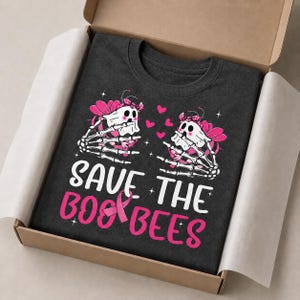 Può includere: Una t-shirt grigio scuro con la grafica di due api fantasma bianche con mani scheletriche, ali rosa e fiocchi rosa. Il testo "SAVE THE BOO BEES" è in bianco e rosa, con un nastro rosa di sensibilizzazione integrato nella lettera B.