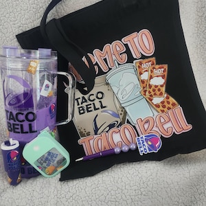 May include: A black tote bag with a Taco Bell design, a clear plastic cup with purple liquid, a small purple pen, a mint green container, and a Taco Bell sticker. The tote bag has the words "Welcome to Taco Bell" printed on it.