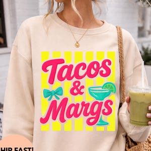 May include: Beige sweatshirt with a vibrant graphic that reads "Tacos & Margs" in pink lettering, set against a yellow striped background. A turquoise bow and margarita glass are also featured. The sweatshirt is a casual, comfortable style.