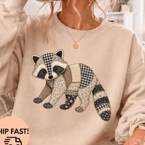 May include: A sand-colored crewneck sweatshirt featuring a central graphic of a raccoon designed with a patchwork aesthetic. The raccoon illustration incorporates various patterns including black and white gingham, floral prints, and solid tan fabric textures.