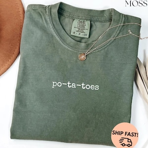 May include: Olive green t-shirt with the word "po-ta-toes" in white script. The shirt has a crew neck and short sleeves. A gold-colored necklace with a pendant rests on the shirt. The shirt is folded on a white surface.