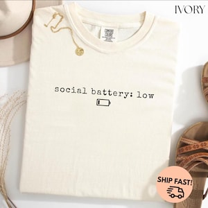以下が含まれることがあります： 「social battery: low」の文字とバッテリーアイコンがプリントされた、アイボリー色のComfort Colors Tシャツ。背景にはゴールドのネックレスとサンダルが見えます。クルーネックのシャツです。