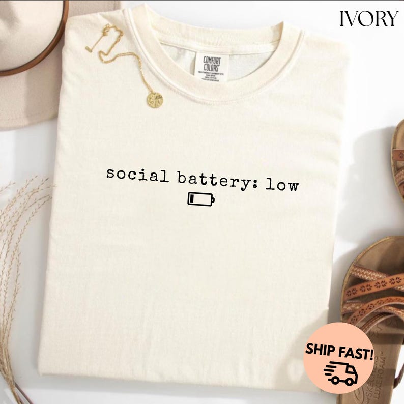 以下が含まれることがあります： 「social battery: low」の文字とバッテリーアイコンがプリントされた、アイボリー色のComfort Colors Tシャツ。背景にはゴールドのネックレスとサンダルが見えます。クルーネックのシャツです。