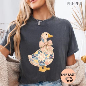 May include: Dark grey t-shirt with a patchwork duck design and a pink bow. The duck has a yellow beak and feet, and the patchwork includes floral and geometric patterns. The word "PEPPER" is printed at the top.