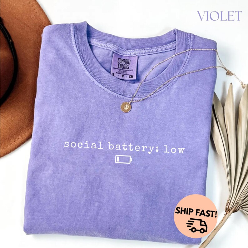 以下が含まれることがあります： 「social battery: low」の文字とバッテリーアイコンがプリントされた、紫色のComfort Colors Tシャツ。 金色のネックレスがTシャツの上に置かれています。 Tシャツは折りたたまれており、背景には茶色の帽子が見えます。