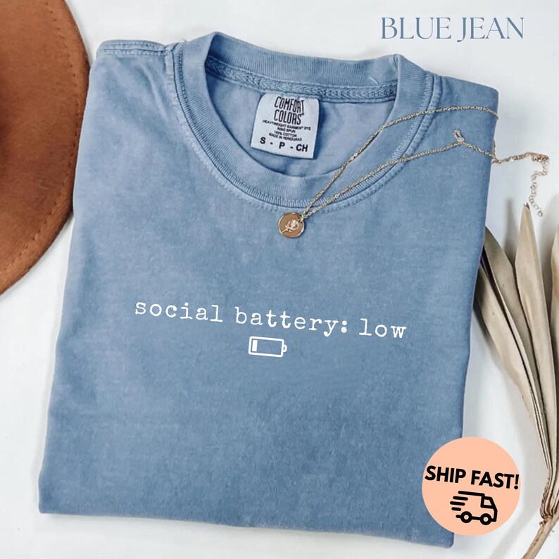 以下が含まれることがあります： 「social battery: low」の文字とバッテリーアイコンが入った、ブルーデニムカラーのTシャツ。ペンダント付きのゴールドネックレスがシャツの上にかけられています。シャツにはComfort Colorsのタグが付いています。