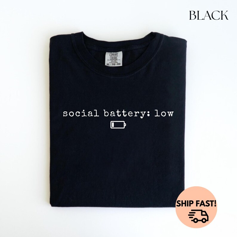 以下が含まれることがあります： 「social battery: low」の白文字とバッテリーアイコンがプリントされた黒いTシャツ。Tシャツは折りたたまれており、上部にタグが付いています。「BLACK」の文字が右上隅に印刷されています。