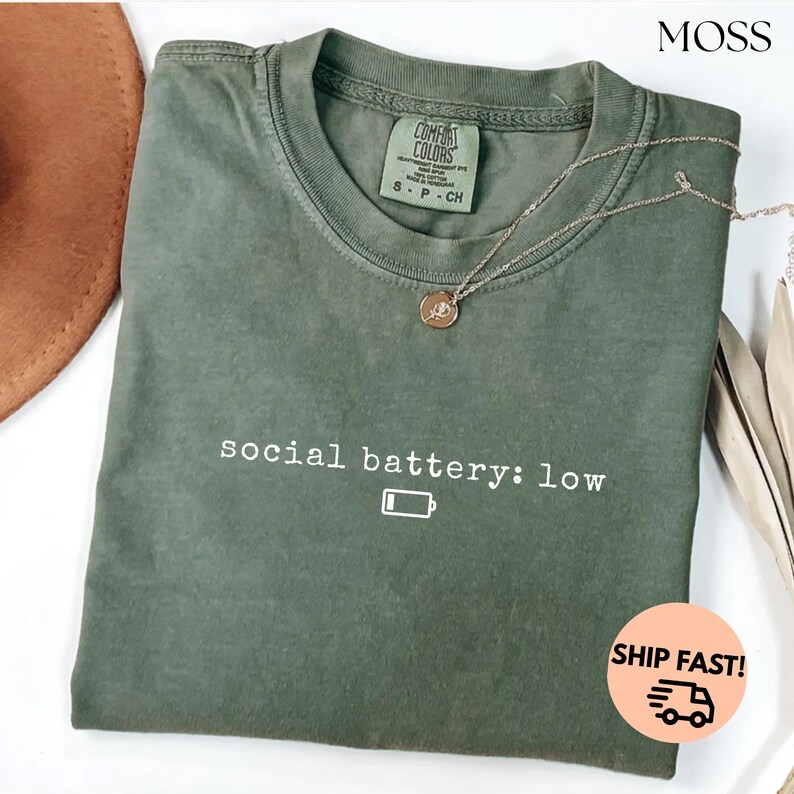 以下が含まれることがあります： 「social battery: low」の文字とバッテリーアイコンがプリントされたオリーブグリーンのTシャツ。 金色のペンダント付きネックレスがシャツの上にかけられています。 シャツにはComfort Colorsのタグが付いています。