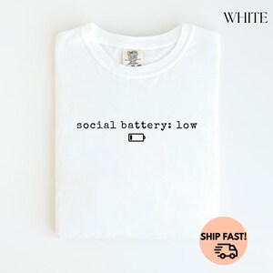 以下が含まれることがあります： 「social battery: low」のテキストと、黒いバッテリー残量アイコンがプリントされた白いTシャツ。Tシャツは折りたたまれ、白い背景に表示されています。右上には黒字で「WHITE」と書かれています。