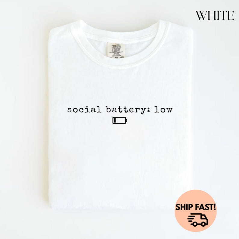 以下が含まれることがあります： 「social battery: low」のテキストと、黒いバッテリー残量アイコンがプリントされた白いTシャツ。Tシャツは折りたたまれ、白い背景に表示されています。右上には黒字で「WHITE」と書かれています。