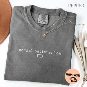 以下が含まれることがあります： 「social battery: low」の文字とバッテリーアイコンが入ったグレーのComfort Colors Tシャツ。シルバーのネックレスがシャツの上に置かれています。シャツは折りたたまれ、白い表面に表示されています。