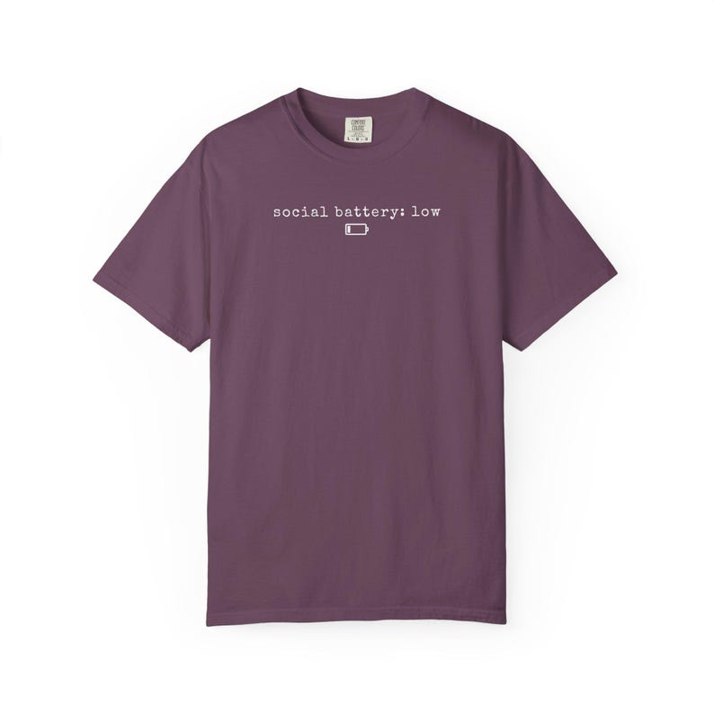 以下が含まれることがあります： 「social battery: low」の文字と白いバッテリーアイコンがプリントされたプラム色のTシャツ。半袖とクルーネックです。生地は柔らかく、着心地の良い素材のようです。