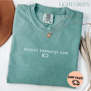 以下が含まれることがあります： 「social battery: low」の文字とバッテリーアイコンがプリントされた、ライトグリーンのTシャツ。クルーネックで半袖です。シルバーのネックレスがTシャツの上にかけられています。