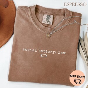 以下が含まれることがあります： 「social battery: low」の文字とバッテリーアイコンが入ったエスプレッソカラーのTシャツ。クルーネックで、Comfort Colorsのタグが付いています。ペンダント付きのシルバーのネックレスがシャツの上にかけられています。