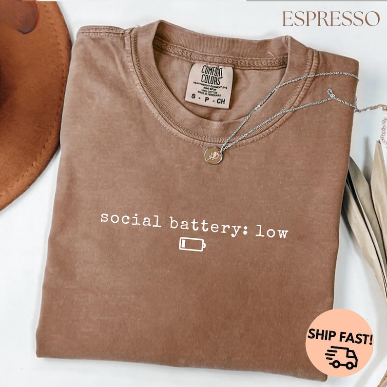 以下が含まれることがあります： 「social battery: low」の文字とバッテリーアイコンが入ったエスプレッソカラーのTシャツ。クルーネックで、Comfort Colorsのタグが付いています。ペンダント付きのシルバーのネックレスがシャツの上にかけられています。
