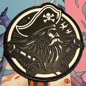 May include: A round, black and white pirate patch featuring a detailed illustration of a pirate with a long beard and a tricorn hat. Two cannons are on either side of the pirate's face, and a skull and crossbones are at the top.