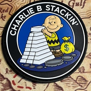 May include: A round black patch with the text "CHARLIE B STACKIN'" in white. The patch features a cartoon character stacking white blocks, with a yellow money bag and coins. The background is blue.