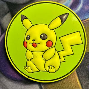 May include: A round, yellow Pikachu character design on a lime green background. The cartoon character has red cheeks, a smiling mouth, and black-tipped ears. The design is set against a colorful, patterned background.
