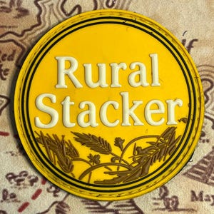 May include: A yellow circular patch with the words "Rural Stacker" in white, outlined in black. The patch features brown wheat stalks and a black border. The background is a map.