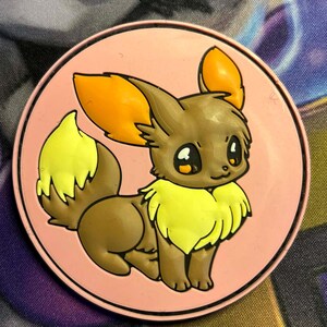 May include: A round, pink Eevee character design. The cartoon Eevee has brown fur, orange ears, and a yellow tail and neck fur. The design is on a pink background.