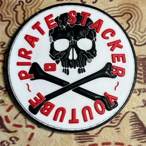 May include: A round, white and black Pirate Stacker YouTube emblem. The emblem features a black skull with red lettering that reads "PIRATE STACKER YOUTUBE". The design includes crossed black bones and a YouTube logo.