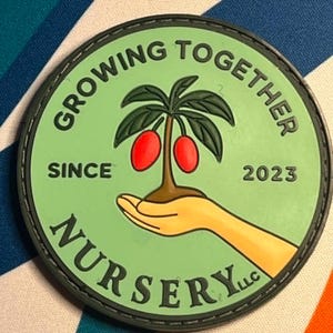 May include: A circular rubber patch with a light green background featuring a graphic of a hand holding a small tree with two red fruits. The text reads "GROWING TOGETHER" at the top, "SINCE 2023" in the middle, and "NURSERY LLC" at the bottom.