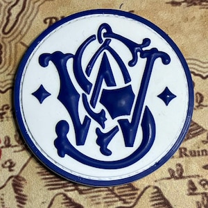 Puede incluir: Un emblema redondo, blanco y azul, con las letras estilizadas "S" y "W" entrelazadas con una "A" central. El emblema tiene un borde azul y está sobre un fondo de mapa. Dos diamantes azules están en el emblema.