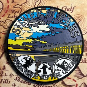 May include: A round, multi-colored medallion with the words "Silver Oceans" and "Stack Wide As The Ocean." The design features a pier, sky, and three circular emblems. The background is a map.