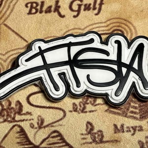 May include: A white and black enamel pin with the word "FISH" in a stylized script. The pin has a black outline and is set against a textured, tan background with the words "Blak Gulf" visible.