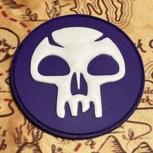 May include: A round, purple patch featuring a white skull design. The skull has large eye sockets and a prominent jawline. The patch is likely an emblem or a decorative element, possibly for clothing or accessories.