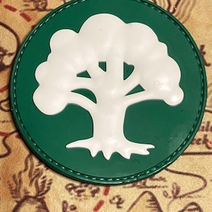 May include: A round, green patch featuring a white tree design. The tree has a detailed trunk and branches, set against a solid green background. The patch is likely an emblem or logo.