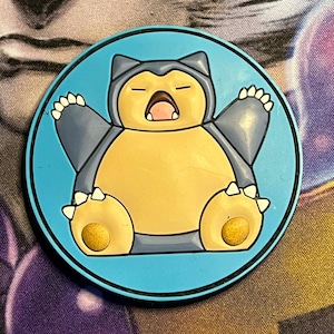 May include: A round, blue and black Pokemon Snorlax design. The Snorlax character is yellow with gray arms and head, and white claws. The character is in a sleeping pose with its mouth open. The design is on a blue background.