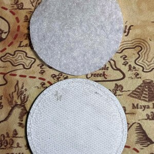 Peut inclure: Deux dessous de verre ronds en tissu gris clair. Le dessous de verre sup&eacute;rieur a une surface textur&eacute;e, tandis que le dessous de verre inf&eacute;rieur a un bord cousu. Le fond pr&eacute;sente un motif de carte dans des tons bruns et beiges.