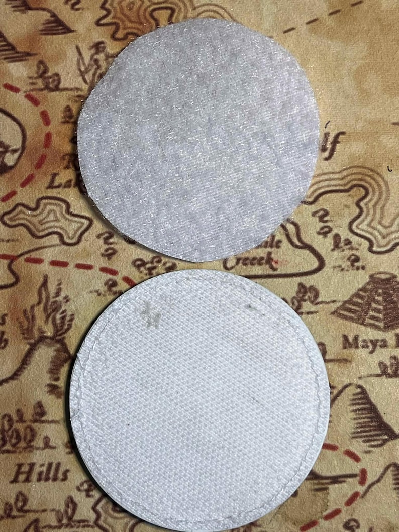 Peut inclure: Deux dessous de verre ronds en tissu gris clair. Le dessous de verre sup&eacute;rieur a une surface textur&eacute;e, tandis que le dessous de verre inf&eacute;rieur a un bord cousu. Le fond pr&eacute;sente un motif de carte dans des tons bruns et beiges.