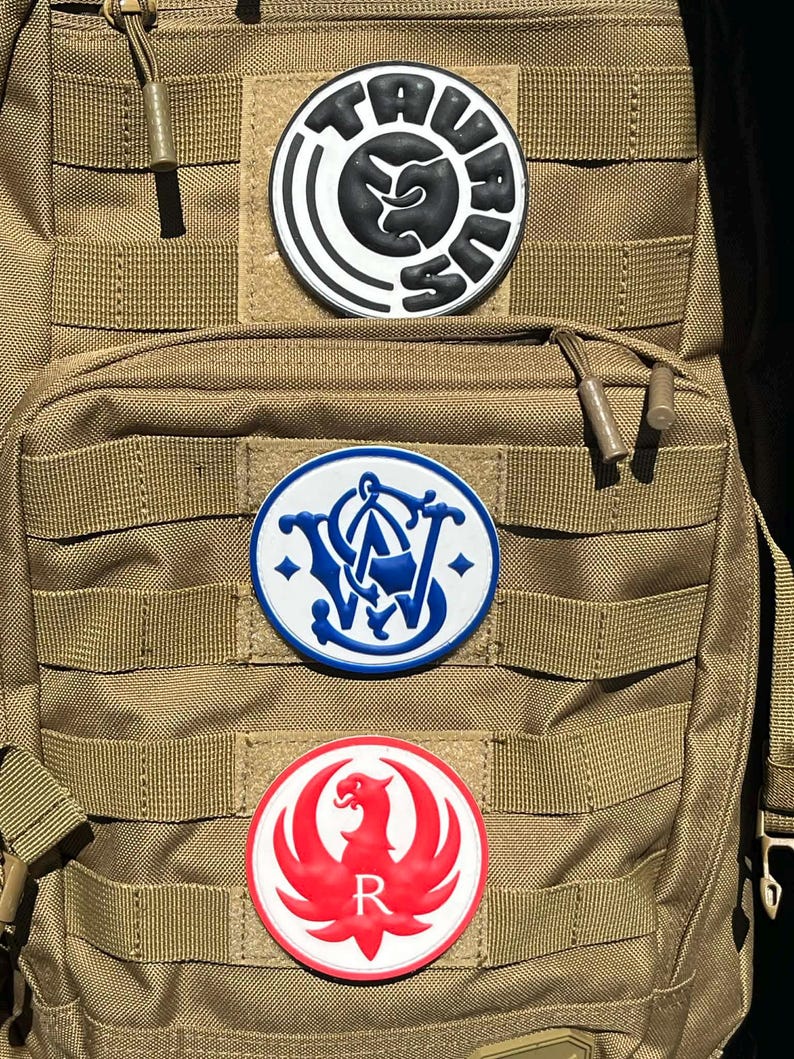 Peut inclure: Un sac &agrave; dos tactique beige avec trois &eacute;cussons circulaires. L'&eacute;cusson sup&eacute;rieur est noir et blanc avec le mot "TAURUS". L'&eacute;cusson du milieu est bleu et blanc avec les lettres "S&W". L'&eacute;cusson inf&eacute;rieur est rouge et blanc avec un ph&eacute;nix et la lettre "R".
