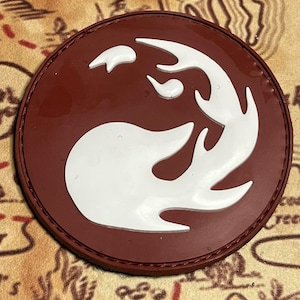 May include: A circular patch with a smooth, dark maroon base and a raised, white, flame-like graphic design centered on the surface. The patch features a stitched border around the circumference, creating a clean, defined edge for the circular shape.