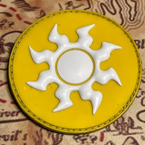 Puede incluir: Un parche redondo amarillo con un diseño de sol blanco. El sol tiene un centro blanco y rayos estilizados. El parche tiene un borde dorado, probablemente un emblema o insignia.