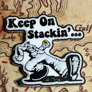 May include: A black and white graphic with the text "Keep On Stackin'" and an illustration of a person. The person is holding a yellow object. The graphic has a black border.